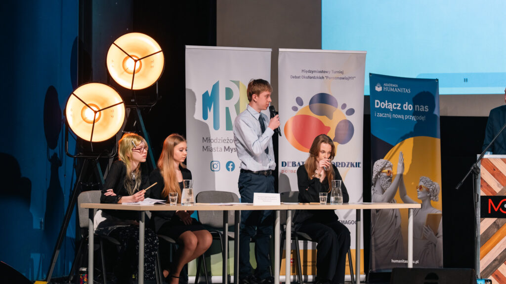 Uczeń przemawia przy stole podczas debaty; pozostali uczestnicy słuchają i notują.