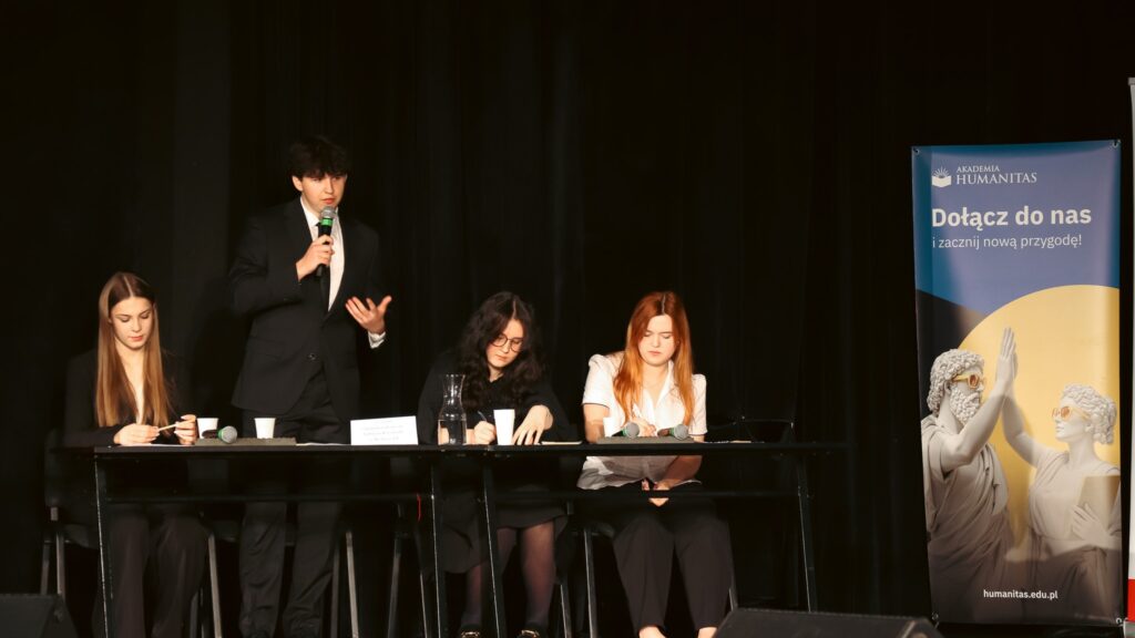 Uczeń przemawia przy stole podczas debaty; pozostali uczestnicy słuchają i notują.