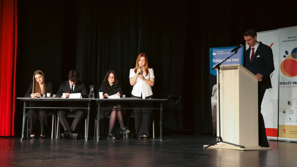 Debata na scenie – jedna osoba przemawia, inni uczestnicy siedzą przy stole, a moderator stoi przy mównicy.