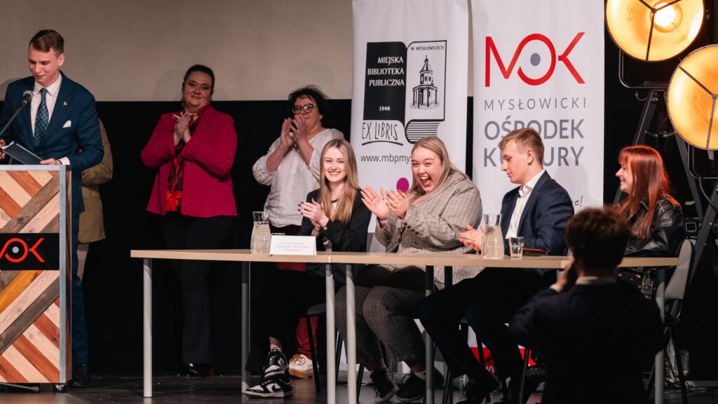 Uczestnicy siedzą przy stole i reagują entuzjastycznie—śmieją się i biją brawo—podczas gdy prowadzący czyta przy mównicy.