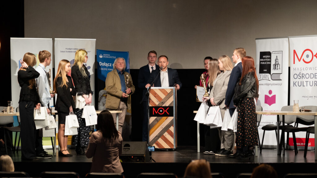 Uczestnicy stoją na scenie i trzymają nagrody oraz torby z upominkami podczas ceremonii wręczenia. Mężczyzna przemawia z mówinicy.