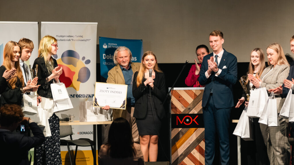 Laureatka przemawia do publiczności, a wokół niej stoją uczestnicy i organizatorzy, bijąc brawo i trzymając nagrody oraz upominki.