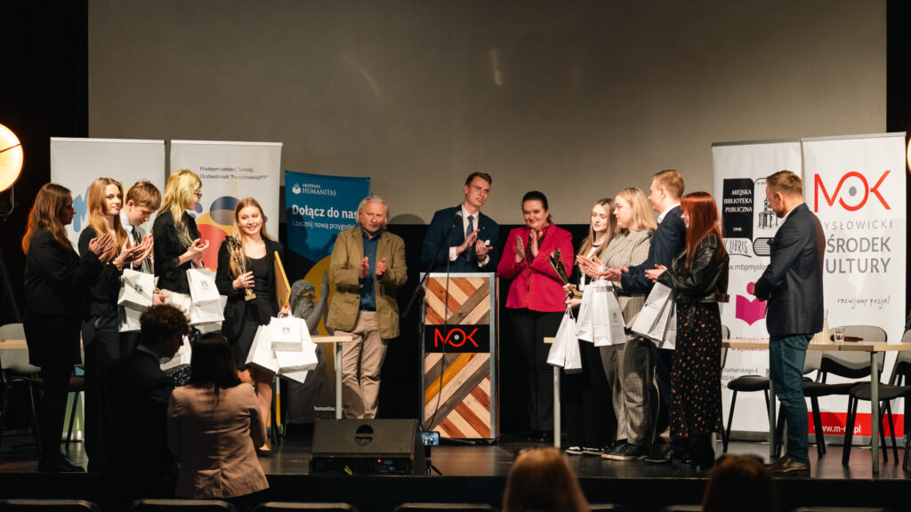 Uczestnicy stoją na scenie, biją brawo i trzymają nagrody oraz torby z upominkami podczas ceremonii wręczenia.