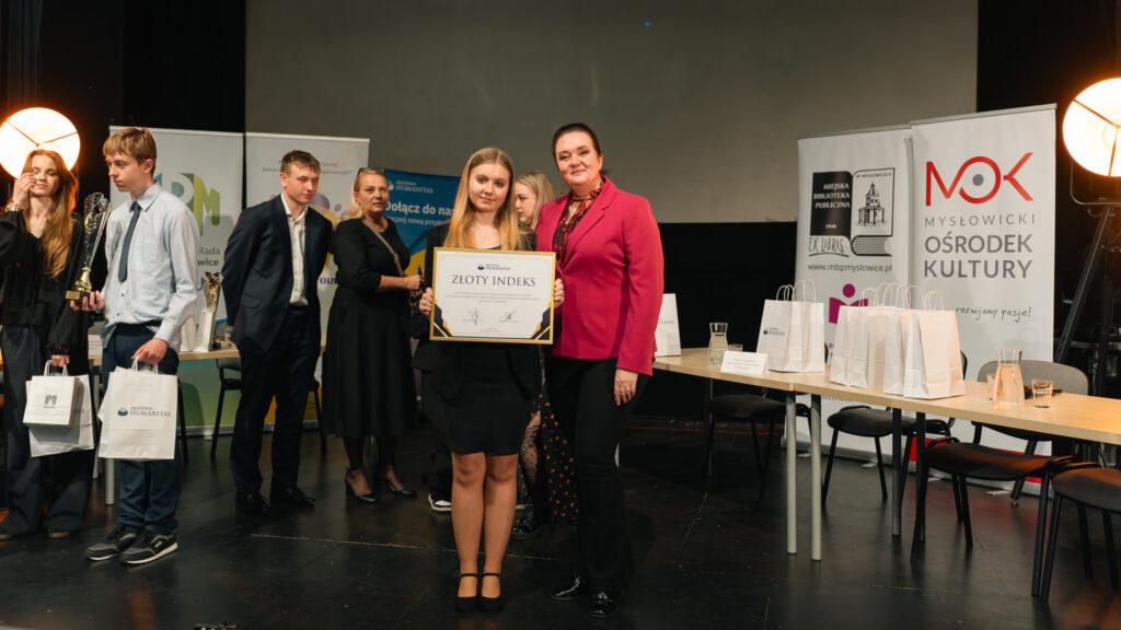 Młoda dziewczyna trzyma dyplom („Złoty Indeks”), stojąc obok kobiety, podczas gdy w tle inni uczestnicy odbierają nagrody i stoją przy stolikach z upominkami.