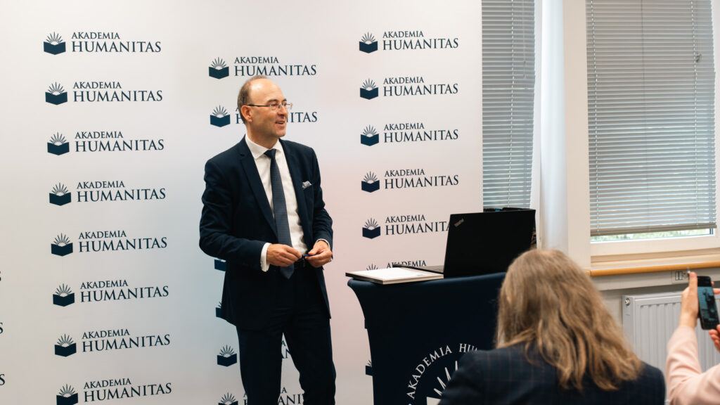 Mężczyzna w garniturze prowadzi wystąpienie w sali Akademii Humanitas, stojąc przy mównicy z laptopem. W tle widoczna jest ścianka z logotypami Akademii Humanitas, a uczestnicy spotkania słuchają prezentacji.