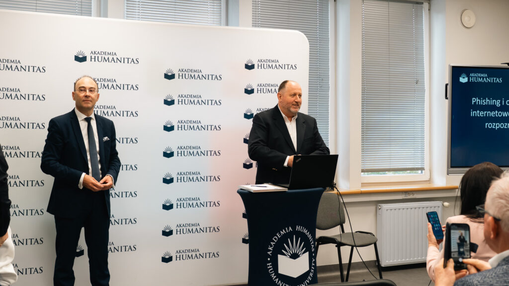 Dwóch mężczyzn prowadzi spotkanie w sali Akademii Humanitas. Jeden stoi przy ściance z logotypami uczelni, drugi przemawia przy mównicy z laptopem, a uczestnicy słuchają i robią zdjęcia telefonami.