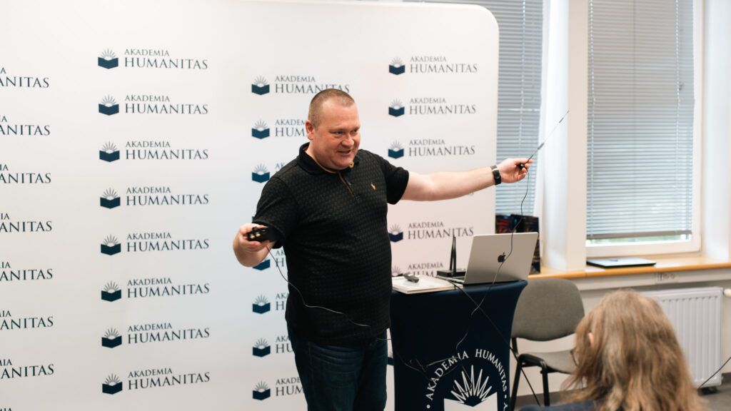 Prelegent w sali Akademii Humanitas prezentuje sprzęt sterujący, trzymając w dłoniach kontroler i antenę podłączone przewodami. W tle widoczna jest ścianka z logotypami Akademii Humanitas oraz stanowisko z laptopem.