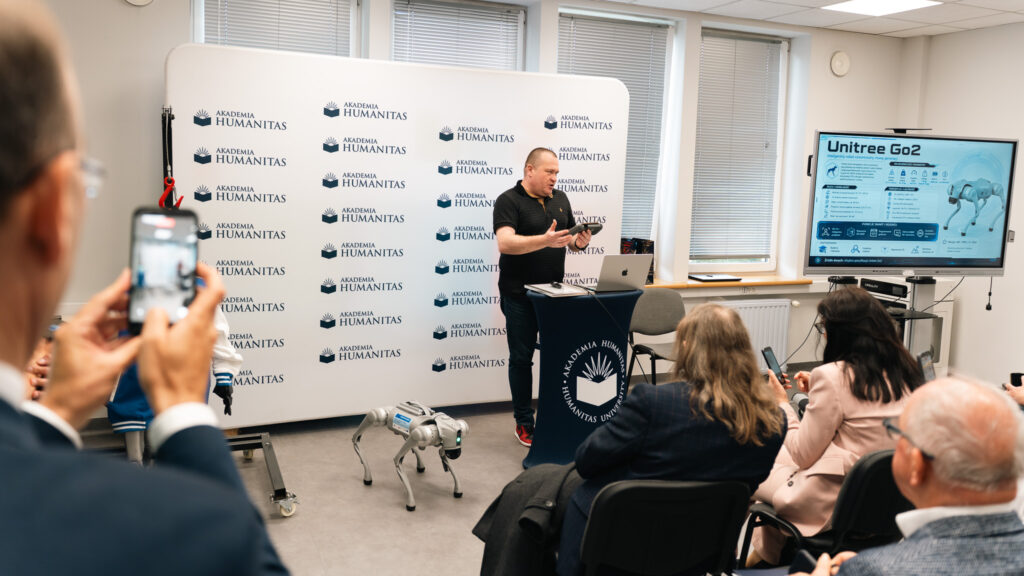 Prelegent prowadzi prezentację w sali Akademii Humanitas, stojąc przy mównicy z logo uczelni. Uczestnicy obserwują pokaz robota Unitree Go2, widocznego na podłodze i na ekranie prezentacyjnym; jedna z osób nagrywa wydarzenie telefonem.