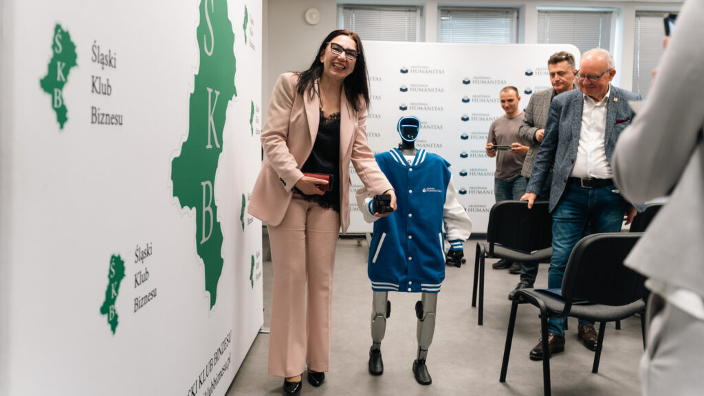 Uczestnicy spotkania w sali Akademii Humanitas obserwują humanoidalnego robota w niebieskiej bluzie uczelni. Obok robota stoi uśmiechnięta kobieta, a w tle widoczne są ścianki z logotypami Akademii Humanitas i Śląskiego Klubu Biznesu.