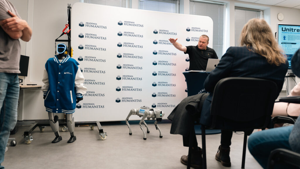 Prelegent prowadzi pokaz robotów w sali Akademii Humanitas. Przed uczestnikami stoją humanoidalny robot w niebieskiej bluzie uczelni oraz robot kroczący, a w tle widoczna jest ścianka z logotypami Akademii Humanitas.