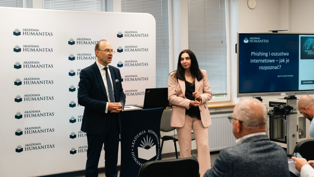 Kobieta i mężczyzna prowadzą spotkanie w sali Akademii Humanitas. W tle widoczna jest ścianka z logotypami uczelni oraz ekran z prezentacją „Phishing i oszustwa internetowe — jak je rozpoznać?”, a uczestnicy słuchają wystąpienia.