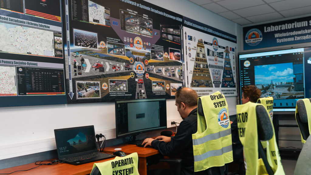Operatorzy systemu pracują przy stanowiskach komputerowych w laboratorium zarządzania kryzysowego. Na ścianach i ekranach widoczne są mapy, panele monitoringu oraz materiały dotyczące systemu SZK Jaśmin, a na krzesłach wiszą kamizelki z napisem „Operator systemu”.