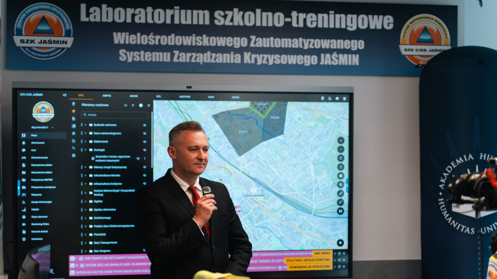 Mężczyzna w garniturze prowadzi wystąpienie w laboratorium szkolno-treningowym systemu zarządzania kryzysowego JAŚMIN. Za nim widoczny jest duży ekran z mapą i panelem systemowym, a obok znajduje się tuba z logo Akademii Humanitas.