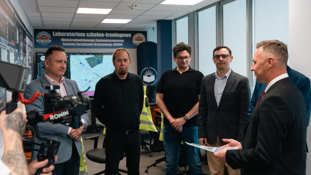 Grupa mężczyzn uczestniczy w spotkaniu w laboratorium szkolno-treningowym systemu zarządzania kryzysowego JAŚMIN. Jeden z prowadzących przemawia, trzymając materiały, a w tle widoczne są ekrany z mapami, stanowiska komputerowe oraz elementy oznakowania Akademii Humanitas.