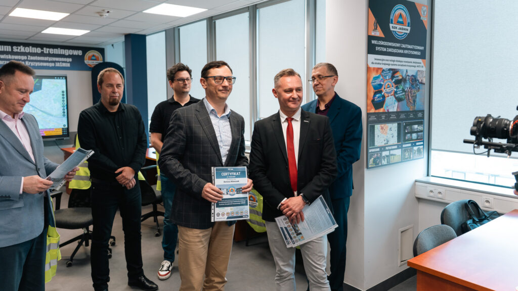 Uczestnicy szkolenia w laboratorium systemu zarządzania kryzysowego JAŚMIN pozują z certyfikatami. W tle widoczne są stanowiska komputerowe, ekran z mapą oraz plakat informacyjny systemu SZK Jaśmin.