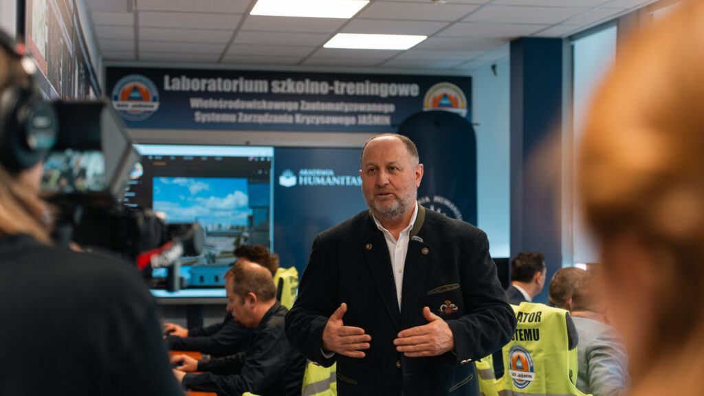 Mężczyzna udziela wypowiedzi w laboratorium szkolno-treningowym systemu zarządzania kryzysowego JAŚMIN. W tle widać osoby pracujące przy stanowiskach komputerowych, ekrany systemowe oraz kamizelki z napisem „Operator systemu”.