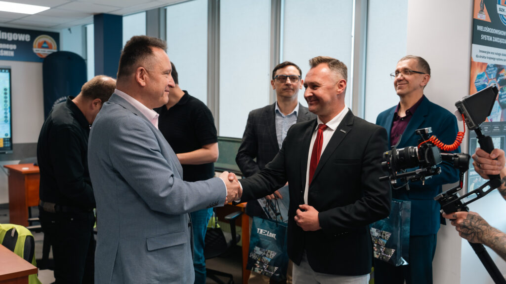 Dwóch mężczyzn ściska sobie dłonie podczas spotkania w laboratorium systemu zarządzania kryzysowego JAŚMIN. Wokół stoją pozostali uczestnicy, a z prawej strony widoczna jest kamera nagrywająca wydarzenie.