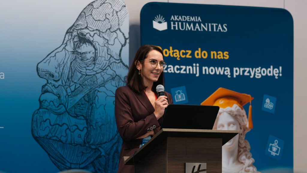 Prelegentka występuje z mikrofonem przy mównicy podczas konferencji Akademii Humanitas. W tle widoczne są banery uczelni z hasłem „Dołącz do nas i zacznij nową przygodę!” oraz grafika związana z konferencją „Medicine in Education. Education in Medicine”.