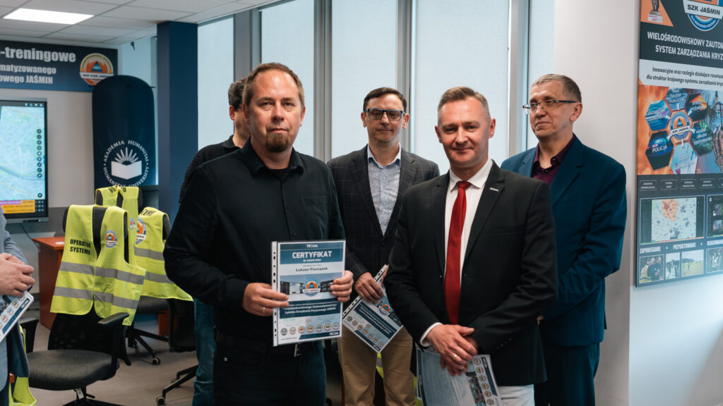 Uczestnicy szkolenia w laboratorium systemu zarządzania kryzysowego JAŚMIN pozują z certyfikatami. Jeden z mężczyzn prezentuje certyfikat, a w tle widoczne są pozostali uczestnicy oraz plakat informacyjny dotyczący systemu SZK Jaśmin.