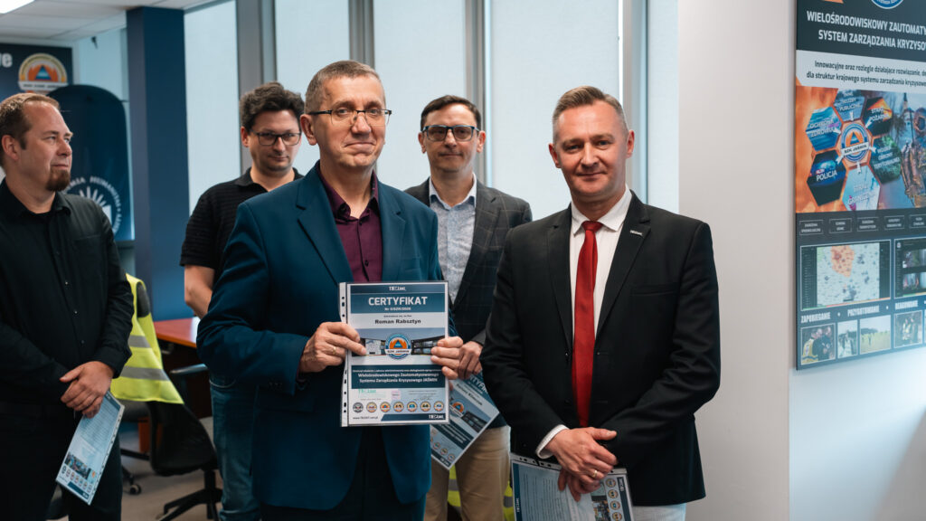 Uczestnicy szkolenia w laboratorium systemu zarządzania kryzysowego JAŚMIN pozują z certyfikatami. Jeden z mężczyzn prezentuje certyfikat, a w tle widoczne są pozostali uczestnicy oraz plakat informacyjny dotyczący systemu SZK Jaśmin.