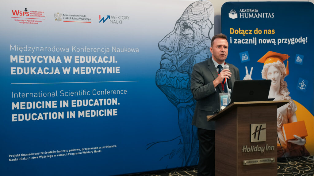 Prelegent występuje z mikrofonem podczas międzynarodowej konferencji naukowej „Medicine in Education. Education in Medicine”. W tle widoczne są ścianka konferencyjna z logotypami organizatorów i informacją o finansowaniu projektu oraz baner Akademii Humanitas z hasłem „Dołącz do nas i zacznij nową przygodę!