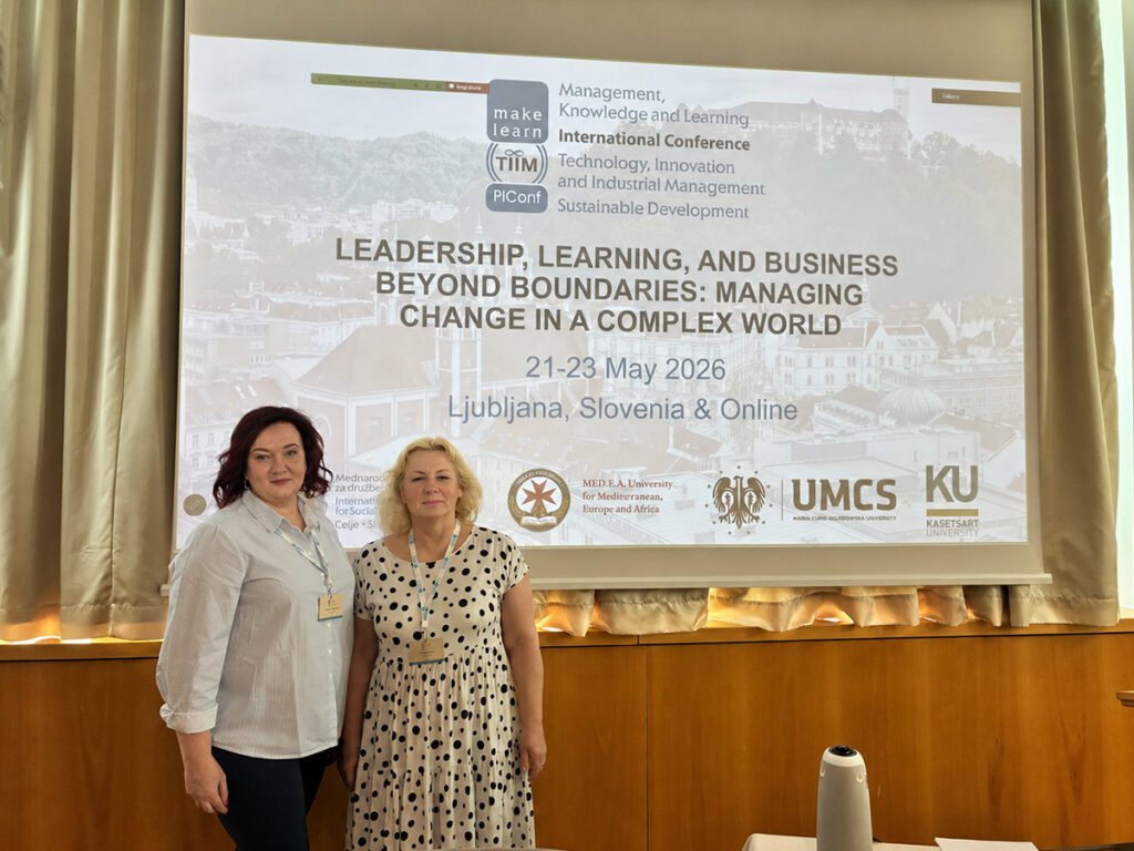 Dwie uczestniczki konferencji stoją przed ekranem z prezentacją wydarzenia „Leadership, Learning and Business Beyond Boundaries: Managing Change in a Complex World”, odbywającego się 21–23 maja 2026 r. w Lublanie i online. Na slajdzie widoczne są logotypy organizatorów oraz partnerów konferencji.