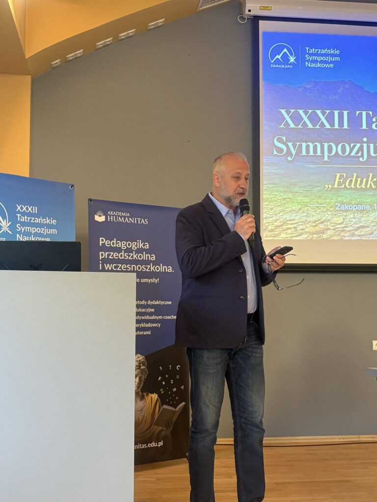 Prelegent przemawia podczas XXXIII Tatrzańskiego Sympozjum Naukowego. Przed nim znajduje się laptop i mikrofon, a w tle widoczny jest baner wydarzenia z górską grafiką.