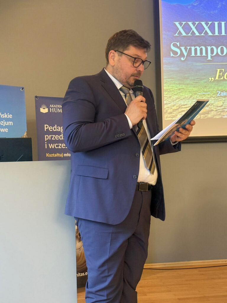 Prelegent przemawia podczas XXXIII Tatrzańskiego Sympozjum Naukowego. Przed nim znajduje się laptop i mikrofon, a w tle widoczny jest baner wydarzenia z górską grafiką.