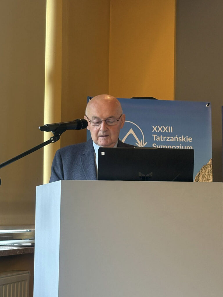 Prelegent przemawia przy mównicy podczas XXXIII Tatrzańskiego Sympozjum Naukowego. Przed nim znajduje się laptop i mikrofon, a w tle widoczny jest baner wydarzenia z górską grafiką.