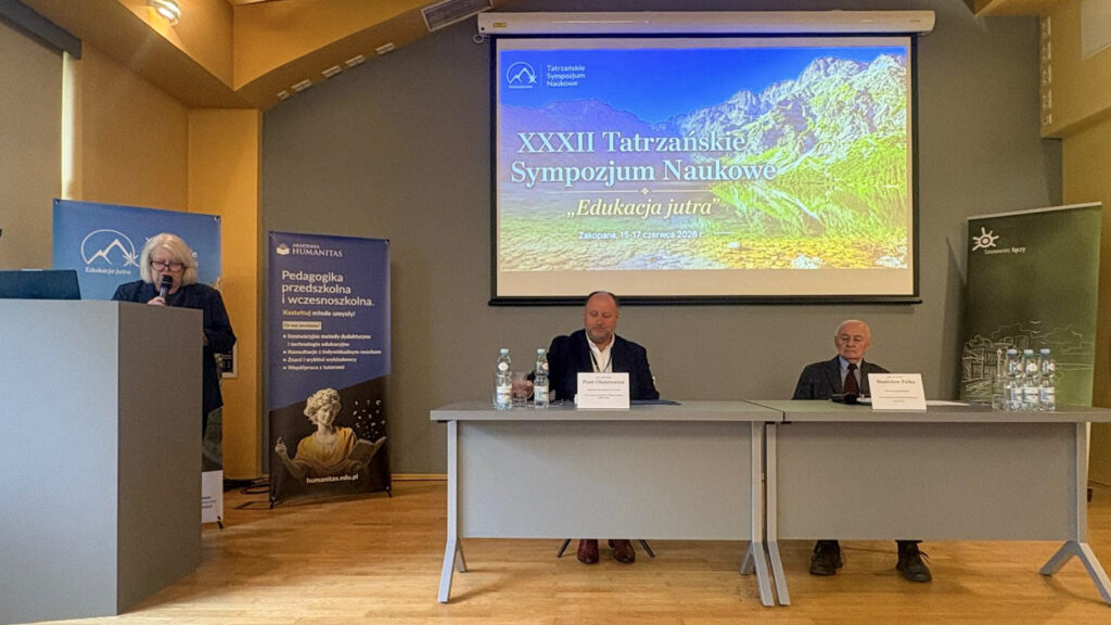 Prelegentka przemawia przy mównicy podczas XXXIII Tatrzańskiego Sympozjum Naukowego „Edukacja Jutra”. Przy stole siedzą paneliści, a na ekranie wyświetlona jest grafika wydarzenia z górskim krajobrazem i datą konferencji w Zakopanem.