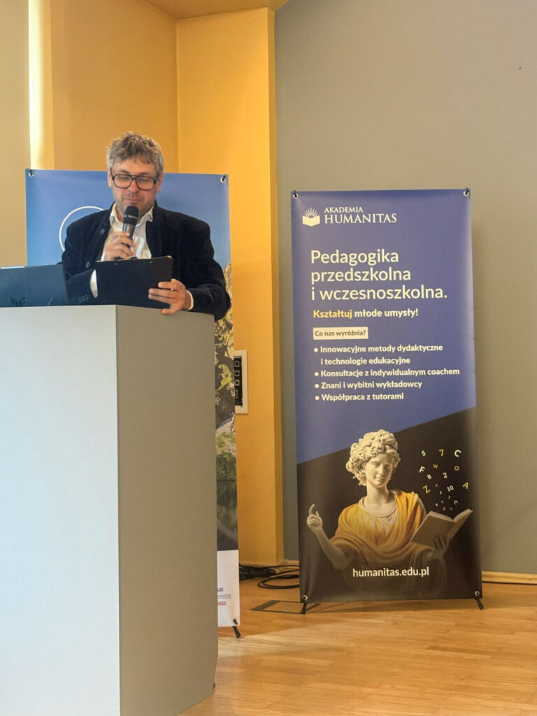 Prelegent przemawia przy mównicy podczas XXXIII Tatrzańskiego Sympozjum Naukowego „Edukacja Jutra”. Trzyma mikrofon, a obok widoczne są banery wydarzenia oraz Akademii Humanitas promujący pedagogikę przedszkolną i wczesnoszkolną.