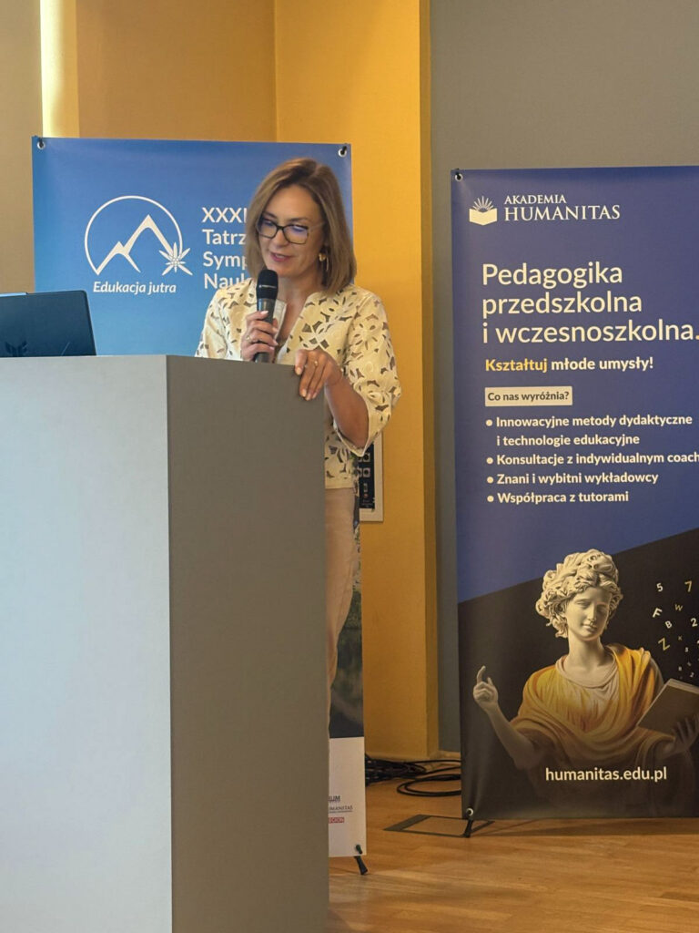Prelegentka przemawia przy mównicy podczas XXXIII Tatrzańskiego Sympozjum Naukowego. Trzyma mikrofon, a obok widoczne są banery wydarzenia „Edukacja Jutra” oraz Akademii Humanitas promujący pedagogikę przedszkolną i wczesnoszkolną.
