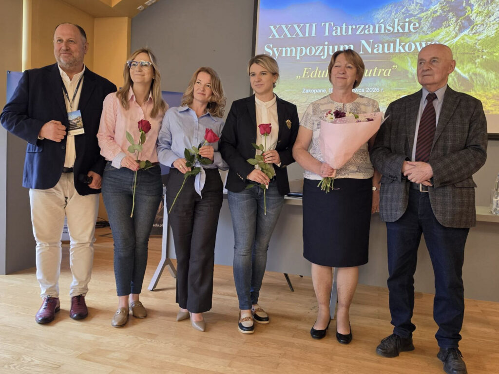 Grupa prelegentów i organizatorów pozuje po wystąpieniu podczas XXXIII Tatrzańskiego Sympozjum Naukowego „Edukacja Jutra” w Zakopanem. Część osób trzyma róże i bukiet kwiatów, a w tle widoczny jest ekran z grafiką wydarzenia.
