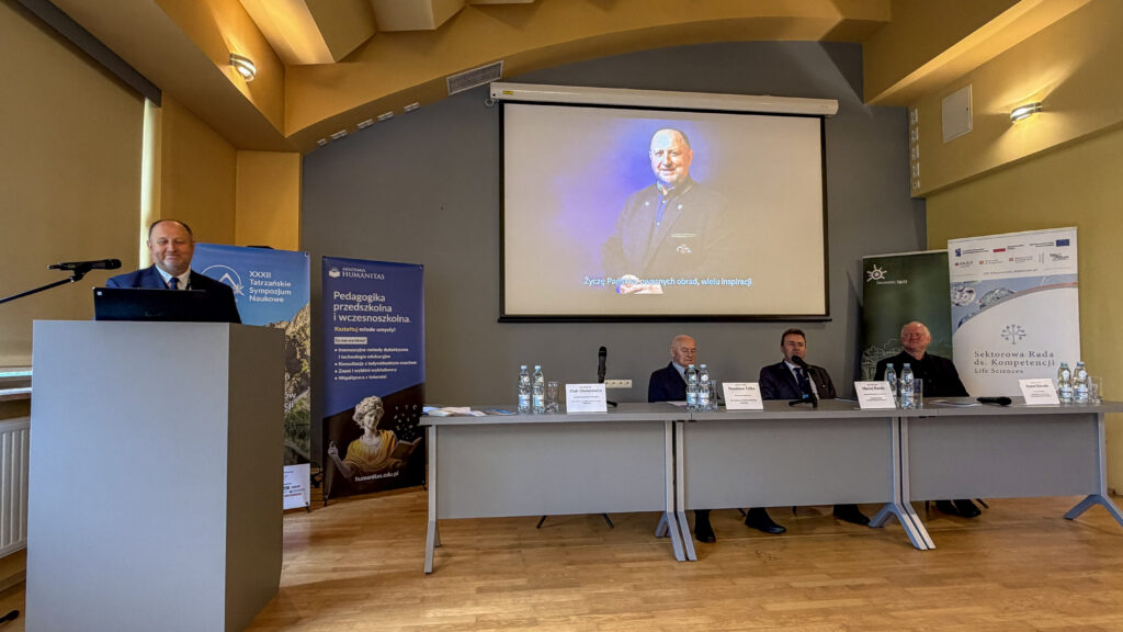 Otwarcie XXXIII Tatrzańskiego Sympozjum Naukowego w sali konferencyjnej. Mężczyzna przemawia przy mównicy, przy stole siedzą paneliści, a na ekranie wyświetlone jest powitanie uczestników; w tle widoczne są banery Akademii Humanitas, sympozjum oraz Sektorowej Rady ds. Kompetencji Life Sciences.