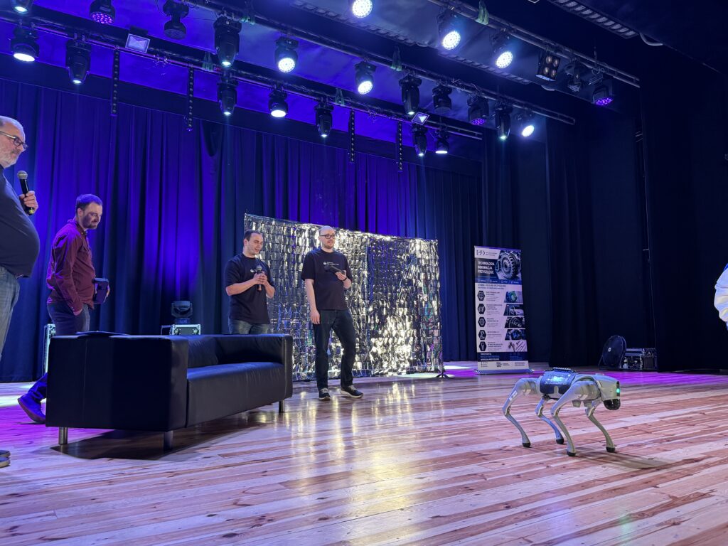 Pokaz robota kroczącego na scenie podczas wydarzenia technologicznego. Kilku prowadzących stoi z mikrofonami, a w tle widoczne są oświetlenie sceniczne, baner dotyczący technologii przyszłości oraz srebrna dekoracja sceny.