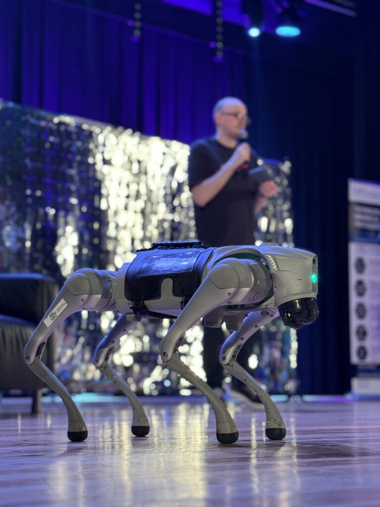 Robot kroczący prezentowany na scenie podczas wydarzenia technologicznego. W tle, poza głębią ostrości, widoczny jest prowadzący z mikrofonem oraz scenografia z ciemną kurtyną, srebrną dekoracją i niebiesko-fioletowym oświetleniem.