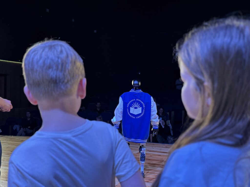 Dzieci obserwują humanoidalnego robota stojącego na scenie tyłem do widowni. Robot ma na sobie niebieską bluzę z logo Akademii Humanitas, a scena jest oświetlona niebieskim światłem.