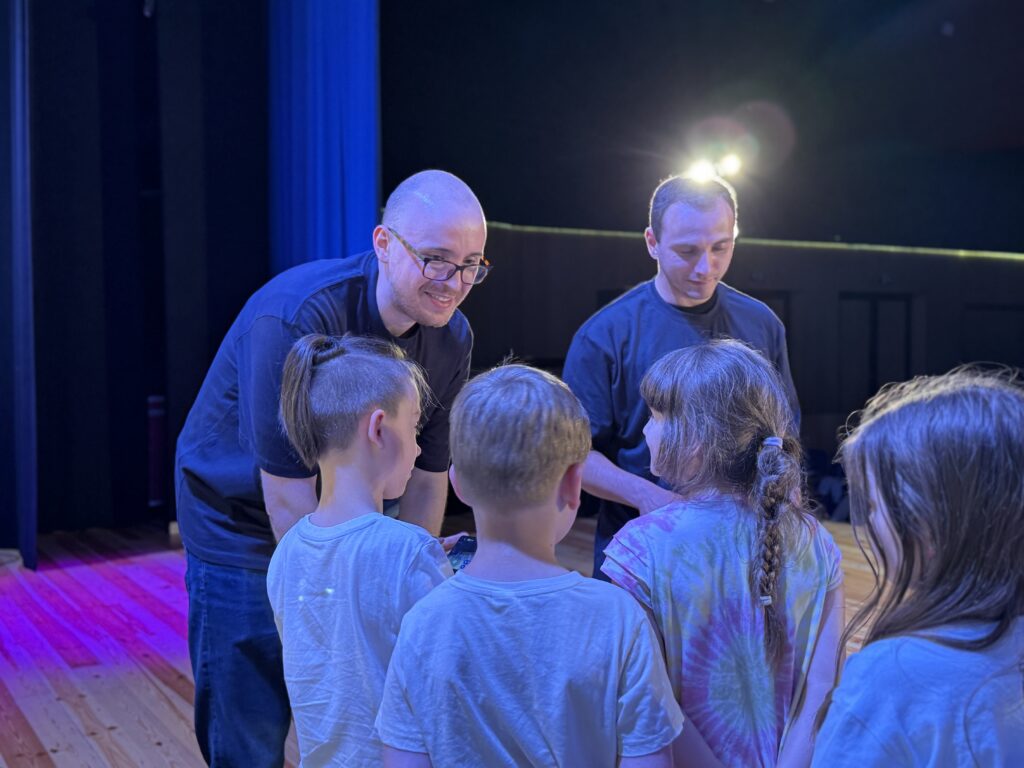 Dwoje prowadzących rozmawia z grupą dzieci na scenie podczas wydarzenia technologicznego. Scena jest oświetlona na niebiesko i fioletowo, a dzieci stoją wokół prowadzących, obserwując pokaz z bliska.