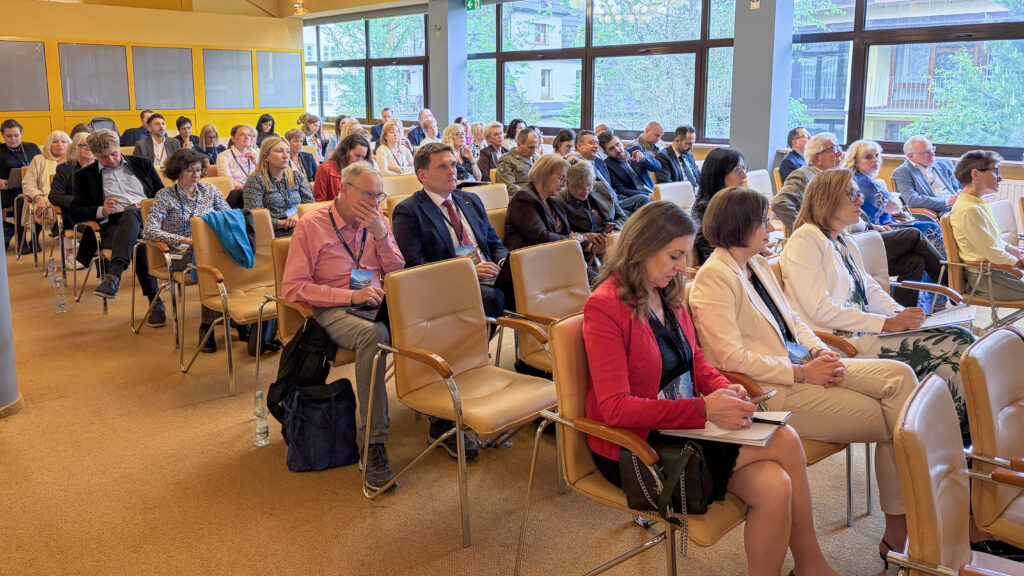 Uczestnicy konferencji siedzą w sali i słuchają wystąpienia. Część osób robi notatki lub pracuje na laptopie, a przez duże okna w tle widać zieleń i budynki za salą.