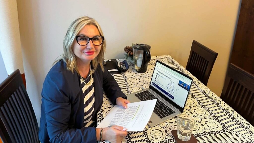 Kobieta uczestniczy w wydarzeniu online, siedząc przy stole z laptopem i notatkami. Na ekranie widoczna jest prezentacja, a obok laptopa znajdują się dokumenty, szklanka wody i czajnik.