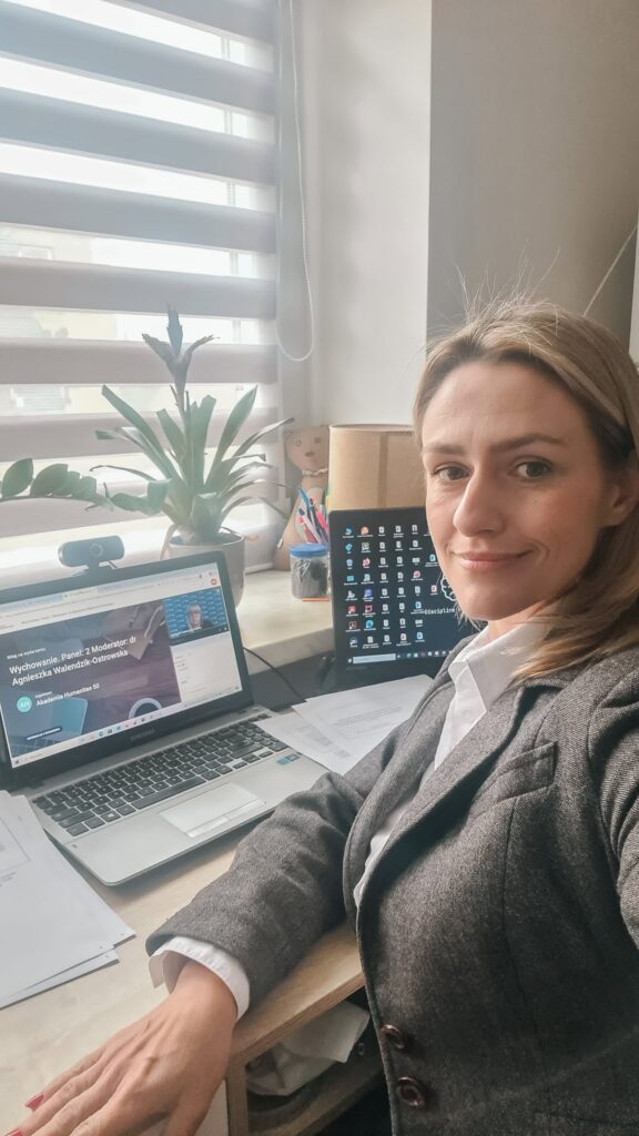 Kobieta siedzi przy biurku i uczestniczy w panelu online Akademii Humanitas na laptopie. Na ekranie widoczna jest wideokonferencja dotycząca wychowania, obok znajduje się drugi ekran z pulpitem komputera, a na biurku leżą dokumenty i stoi roślina.