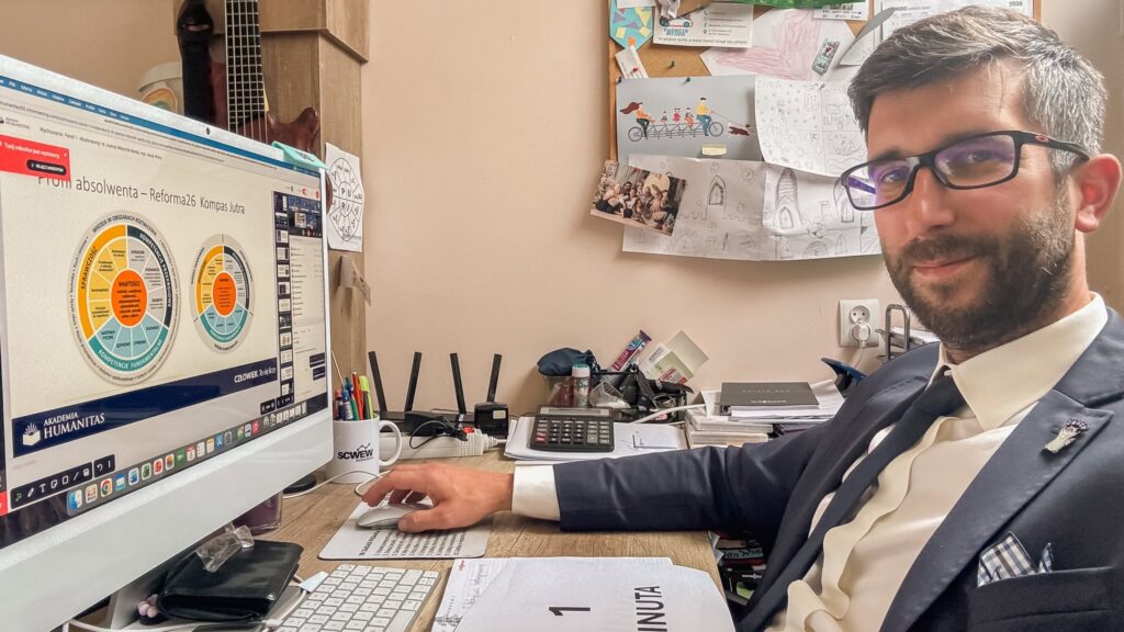 Mężczyzna w garniturze uczestniczy w panelu online Akademii Humanitas, siedząc przy komputerze w gabinecie. Na ekranie widoczna jest prezentacja „Profil absolwenta – Reforma26 Kompas Jutra” oraz okno wideokonferencji z uczestnikami spotkania.