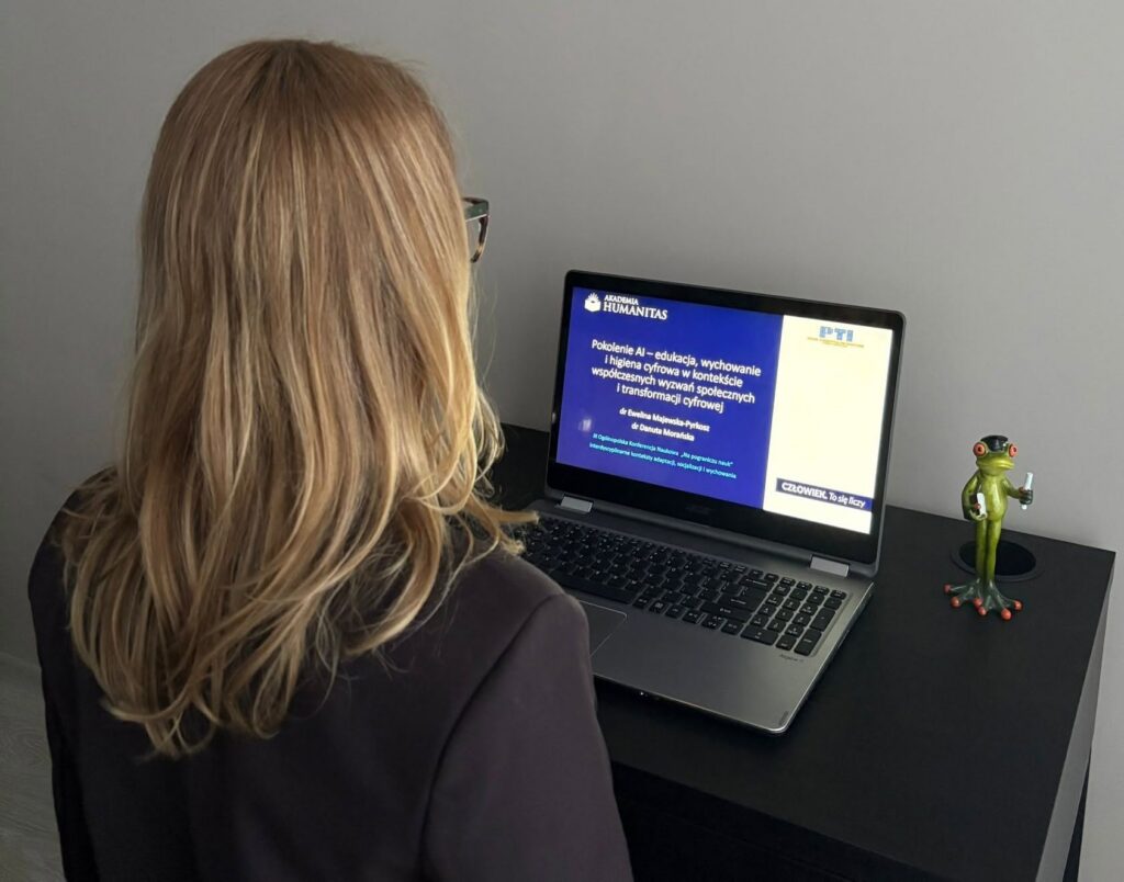 Kobieta ogląda prowadzi online Akademii Humanitas na laptopie. Na ekranie widoczny jest slajd dotyczący pokolenia AI, edukacji, wychowania i higieny cyfrowej w kontekście współczesnych wyzwań społecznych, a obok laptopa stoi mała figurka żaby.