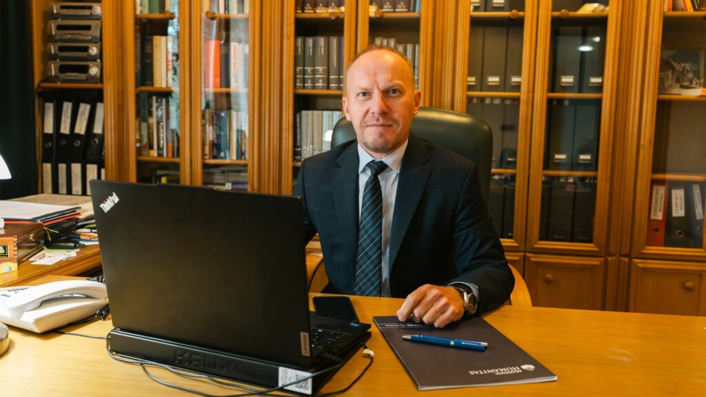Rektor Akademii Humanitas, dr Mariusz Lekston siedzi przy biurku w gabinecie, przed otwartym laptopem. W tle widoczne są drewniane regały z książkami i dokumentami, a na biurku leżą materiały Akademii Humanitas, telefon oraz długopis.