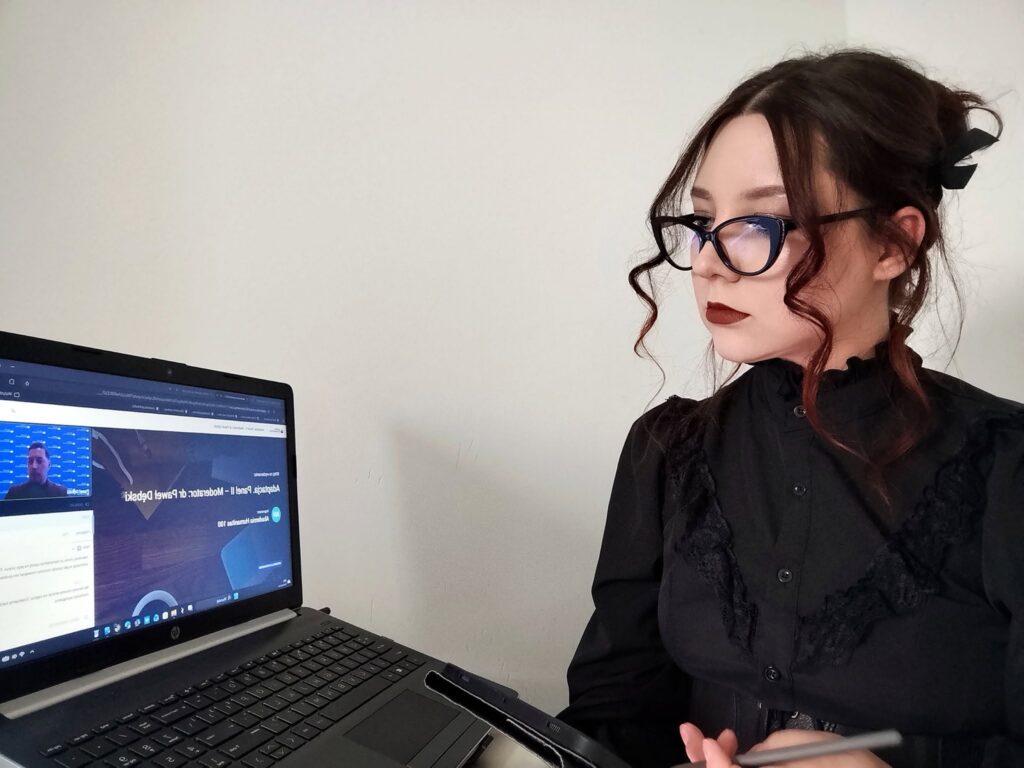 Studentka uczestniczy w zajęciach online, siedząc przy laptopie z uruchomioną wideokonferencją. Trzyma tablet graficzny i rysik, a na ekranie widoczna jest prezentacja oraz prowadzący spotkanie.