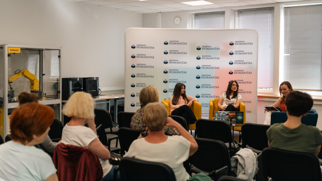 Panel dyskusyjny w sali Akademii Humanitas z udziałem trzech prelegentek siedzących na fotelach przed ścianką z logotypami uczelni. Na widowni siedzą uczestnicy spotkania, a po lewej stronie sali widoczne jest stanowisko z robotem przemysłowym.