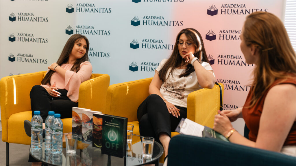 Trzy kobiety prowadzą panel dyskusyjny w sali Akademii Humanitas, siedząc na żółtych fotelach przed ścianką z logotypami uczelni. Na stoliku widoczne są książki i woda, a uczestnicy spotkania słuchają rozmowy z widowni.