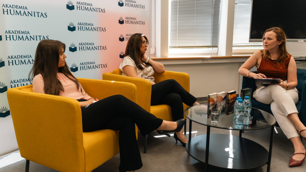 Trzy kobiety prowadzą panel dyskusyjny w sali Akademii Humanitas, siedząc na żółtych fotelach przed ścianką z logotypami uczelni. Na stoliku widoczne są książki i woda, a uczestnicy spotkania słuchają rozmowy z widowni.