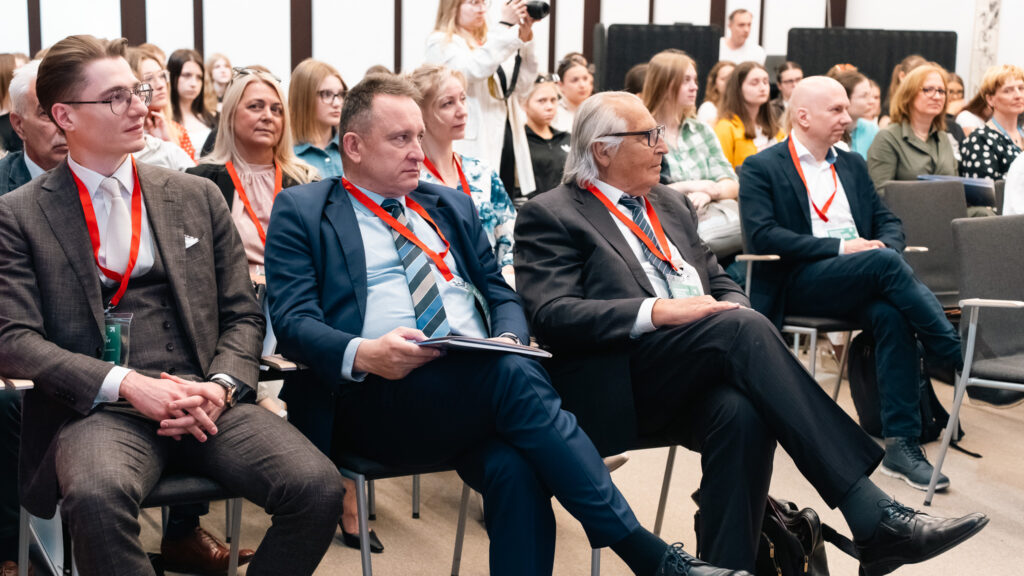 Uczestnicy konferencji siedzą w sali i słuchają wystąpienia. W pierwszym rzędzie widoczni są mężczyźni w garniturach z identyfikatorami, a za nimi kolejne osoby biorące udział w wydarzeniu.