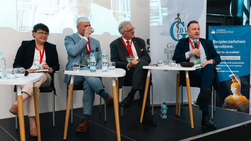 Panel dyskusyjny podczas konferencji dotyczącej bezpieczeństwa i ratownictwa medycznego. Czterech prelegentów siedzi przy stolikach na scenie, jeden z nich mówi do mikrofonu, a w tle widoczne są logo Głównej Biblioteki Lekarskiej oraz baner Akademii Humanitas.